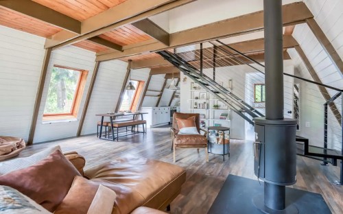 Cozy interior of a modern cabin with wooden beams, a staircase, and large windows letting in natural light.
