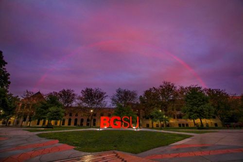 A vibrant rainbow arches over the BGSU sign, surrounded by trees and a cloudy pink sky at dusk.
