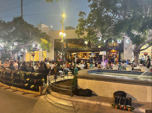 Outdoor dining scene at night with people gathered around tables, trees with lights, and a fountain in the foreground.