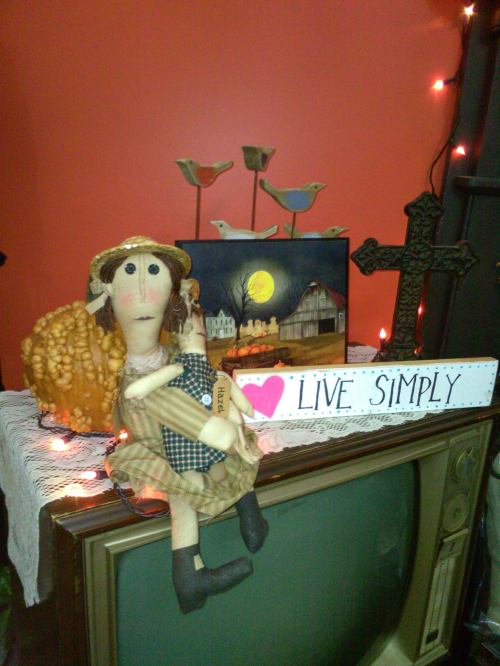 A decorative display featuring a doll, a sign that says "Live Simply," and various ornaments on a vintage TV.