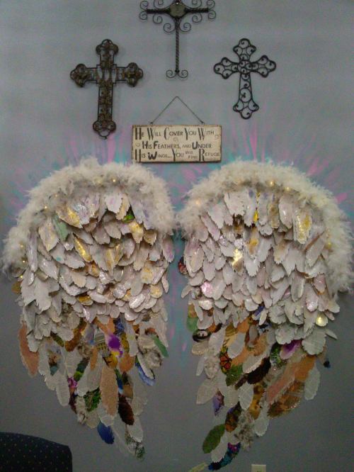 Decorative angel wings made of feathers and colorful materials, displayed against a wall with crosses and a sign.