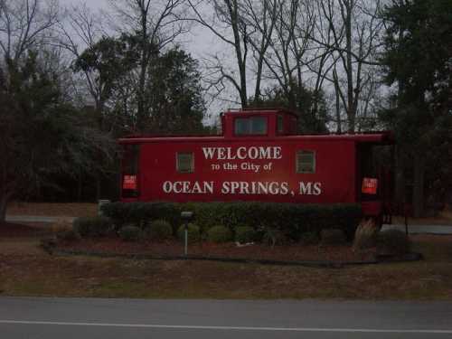 A red caboose sign welcoming visitors to Ocean Springs, MS, surrounded by greenery and trees.