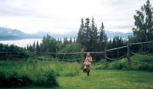 A woman walks through a lush green field with mountains and trees in the background under a cloudy sky.