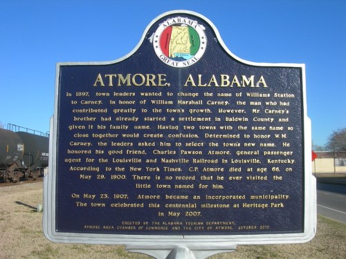 Sign about Atmore, Alabama's history, including its name change and notable figures, with a background of train cars.