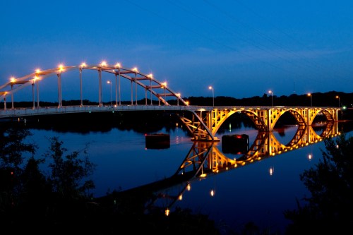A brightly lit bridge arches over a calm river at dusk, reflecting its lights in the water below.