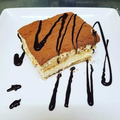 A slice of tiramisu on a white plate, drizzled with chocolate sauce and topped with cocoa powder.