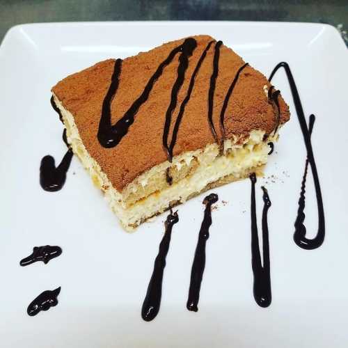 A slice of tiramisu on a white plate, drizzled with chocolate sauce and topped with cocoa powder.