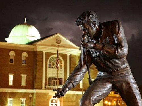 A bronze statue of a singer holding a microphone, with a historic building and cloudy night sky in the background.