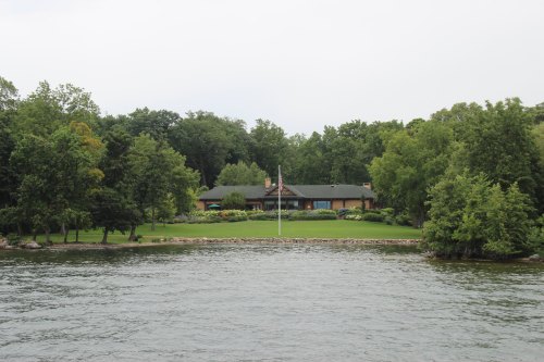 A serene lakeside view of a house surrounded by lush trees and a grassy area, with a flagpole in the foreground.