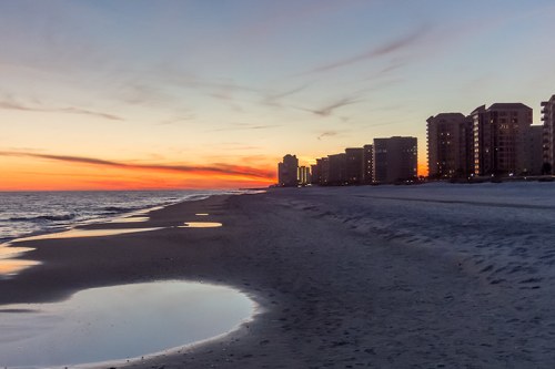 Sunset over a beach with calm waves, reflecting light, and buildings lining the shore in the distance.