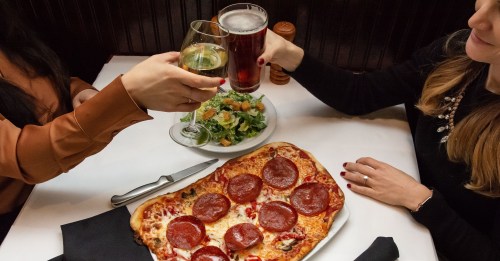 Two people toast with drinks over a table featuring a pepperoni pizza and a salad.