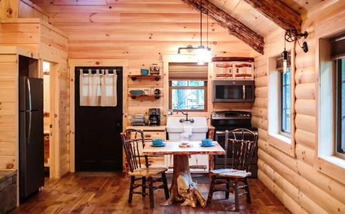 Cozy log cabin kitchen with wooden walls, a dining table, chairs, and modern appliances. Warm, inviting atmosphere.