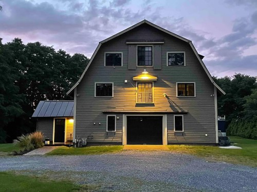 A large, modern barn-style house illuminated at dusk, surrounded by trees and a gravel driveway.