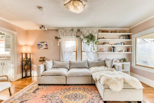 Cozy living room with a light gray sectional sofa, a patterned rug, bookshelves, and plants in a warm, inviting space.