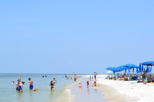 A sunny beach scene with people swimming and relaxing under blue umbrellas along the shore.