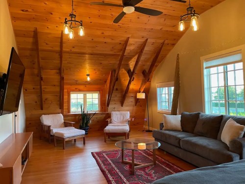 Cozy living room with wooden walls, a sofa, armchairs, a coffee table, and large windows letting in natural light.