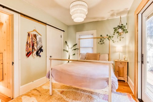 Cozy bedroom with a light wood bed, soft bedding, plants, and a warm light fixture, featuring a rug and natural light.