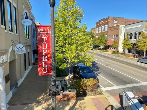 A street view in Tupelo featuring a "Fall into Tupelo" banner and trees lining the sidewalk.