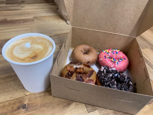 A box of four donuts, including a pink frosted donut with sprinkles, next to a cup of coffee on a wooden table.