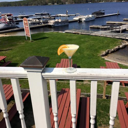 A cocktail sits on a railing with a view of a lake and boats in the background, surrounded by green grass and wooden docks.