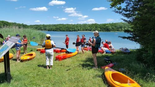 A group of people in life jackets gather by a lake, preparing for kayaking on a sunny day. Kayaks are lined up on the shore.
