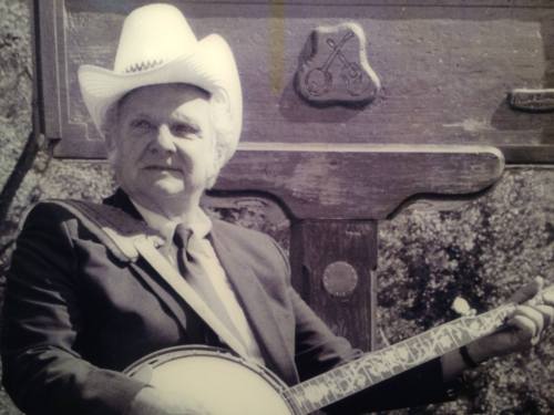 A man in a cowboy hat and suit holds a banjo, smiling in front of a wooden backdrop.