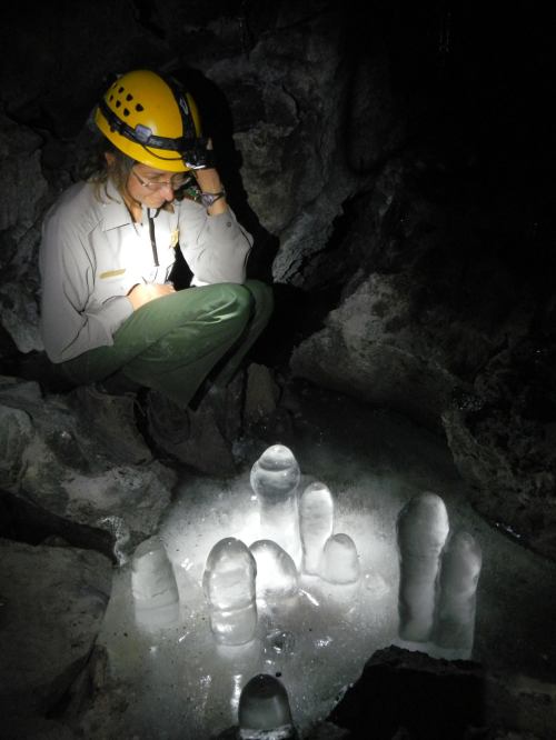 A person in a helmet examines ice formations in a dark cave, illuminated by a light source.