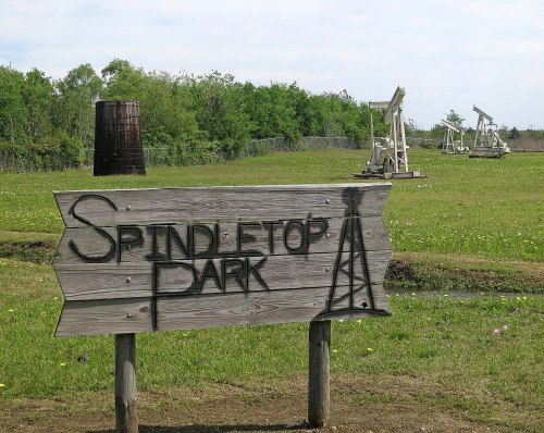 Sign for Spindletop Park with oil derricks in the background, surrounded by green grass and trees under a blue sky.