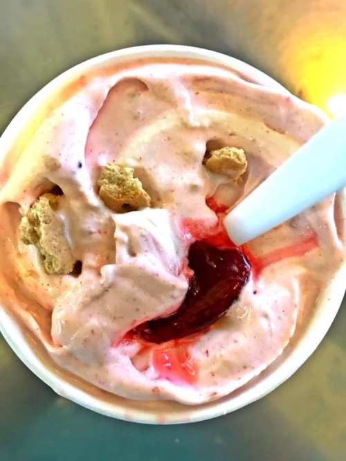 A cup of pink ice cream with a swirl of red sauce and cookie pieces on top, served with a white spoon.