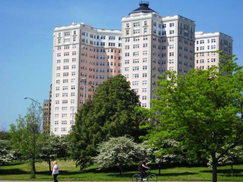 A tall, pink apartment building surrounded by green trees and people walking and biking in a sunny park.