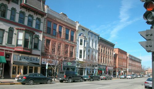A street lined with historic buildings and shops, featuring parked cars and a clear blue sky.
