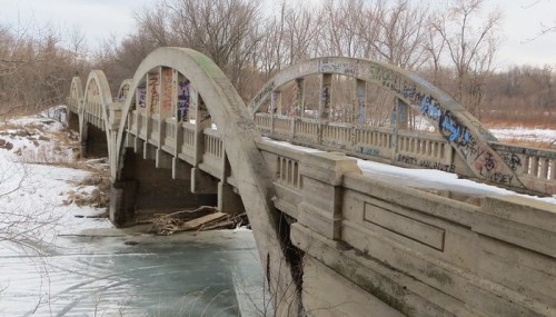 A weathered concrete bridge with arches, covered in graffiti, spans a frozen river surrounded by bare trees.