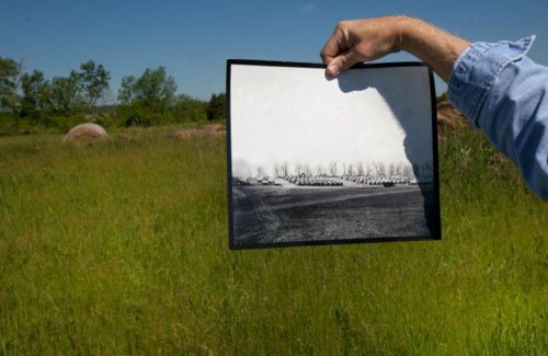 A hand holds a black-and-white photo of a landscape, contrasting with a vibrant green field in the background.