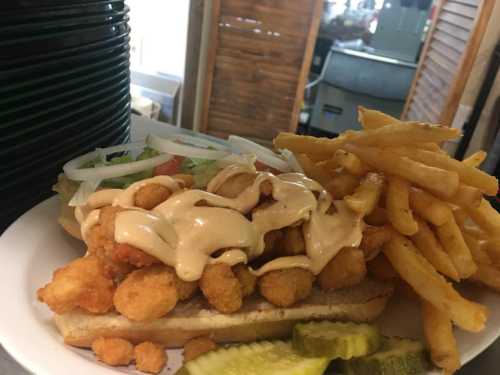 A plate featuring a shrimp sandwich topped with sauce, lettuce, and tomatoes, alongside a serving of fries and pickles.