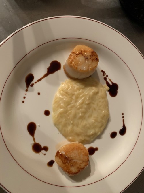 Two seared scallops on a plate with creamy risotto and drizzles of sauce.