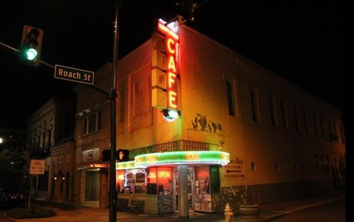 A brightly lit café at night, featuring neon signs and a corner street sign for Roach St.