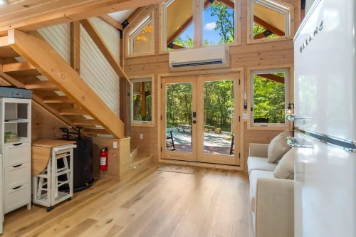 Cozy wooden interior of a tiny house with large windows, a staircase, and a view of the outdoors.