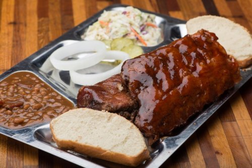 A metal tray with BBQ ribs, coleslaw, baked beans, pickles, onion slices, and two slices of bread.