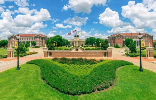A sunny view of the University of Southern Mississippi campus, featuring green lawns, buildings, and a blue sky with clouds.
