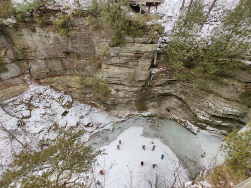 A snowy canyon with steep rock walls, featuring a frozen pond and small figures exploring the icy landscape below.