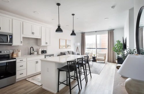 Modern kitchen and living area with white cabinets, black accents, a dining table, and large windows letting in natural light.