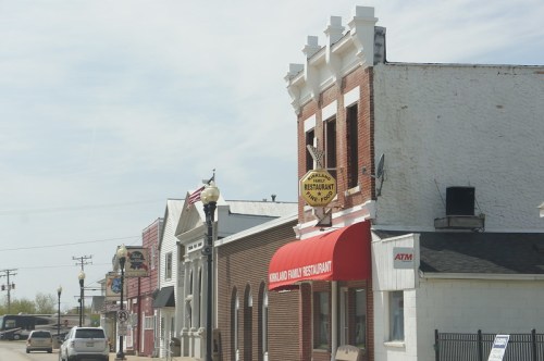 A small-town street featuring a restaurant with a red awning and vintage signage, alongside other quaint buildings.