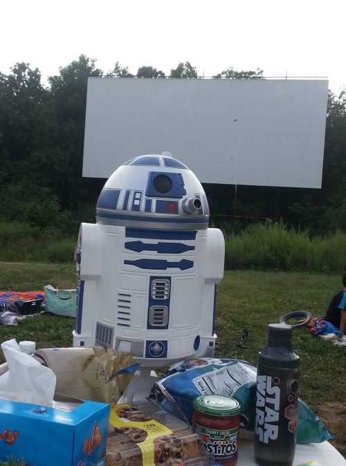 A life-sized R2-D2 figure in front of a blank outdoor movie screen, surrounded by snacks and drinks.