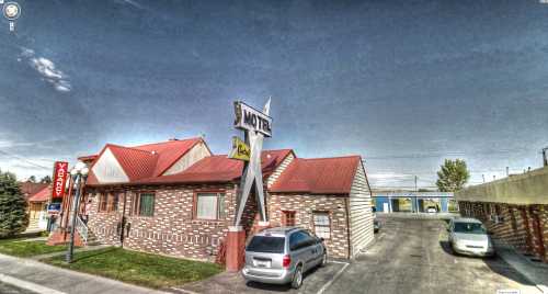 A brick motel with a red roof and a "Vacancy" sign, surrounded by parked cars and a clear blue sky.