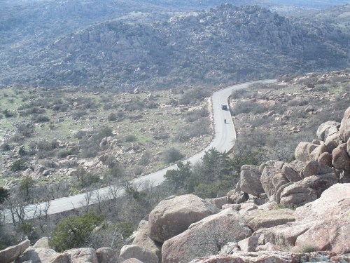A winding road curves through rocky terrain and sparse vegetation, with hills in the background under a clear sky.