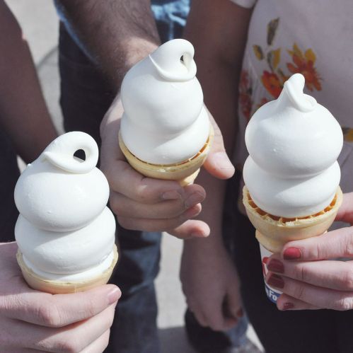 Three hands holding ice cream cones with soft serve, topped with a swirl, against a blurred background.