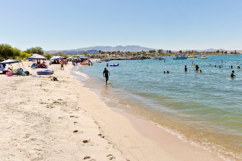 A sunny beach scene with people swimming, lounging under umbrellas, and a person walking along the shore.