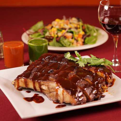 A plate of glazed ribs with sauce, garnished with cilantro, alongside a colorful salad and glasses of wine.