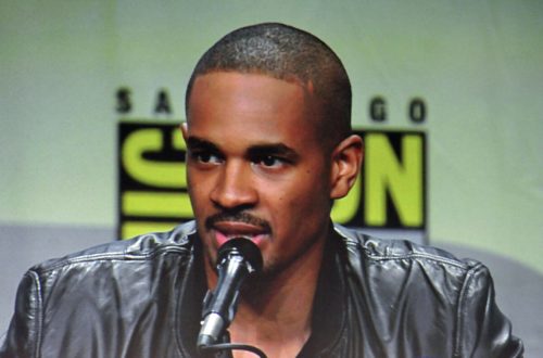 A man in a black leather jacket speaks into a microphone at a panel event, with a San Diego Comic-Con backdrop.