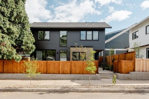 Modern two-story house with dark siding, large windows, and a wooden fence, surrounded by greenery and a clear blue sky.